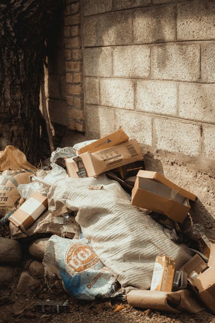 A large collection of mixed waste materials is piled up on a paved sidewalk in front of a commercial building, with various types of rubbish including cardboard boxes, plastic bags, paper, and packagi