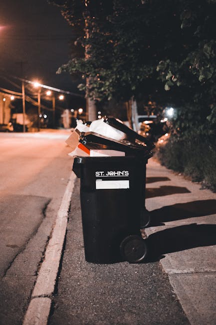 A black wheelie bin with a white label and the name 'ST. JOHN'S' printed on the front is positioned on the edge of a residential pavement at night. The bin is filled with various discarded items, incl