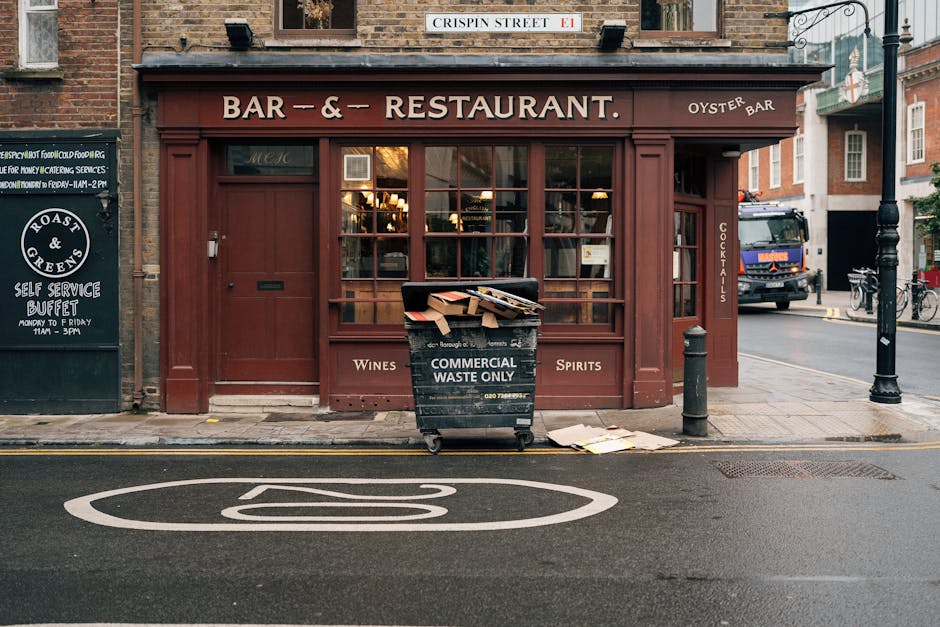 On the sidewalk in front of a restaurant with a deep red, wooden exterior, a black wheeled bin labeled 'COMMERCIAL WASTE ONLY' is filled with flattened cardboard boxes and paper debris, some of which 